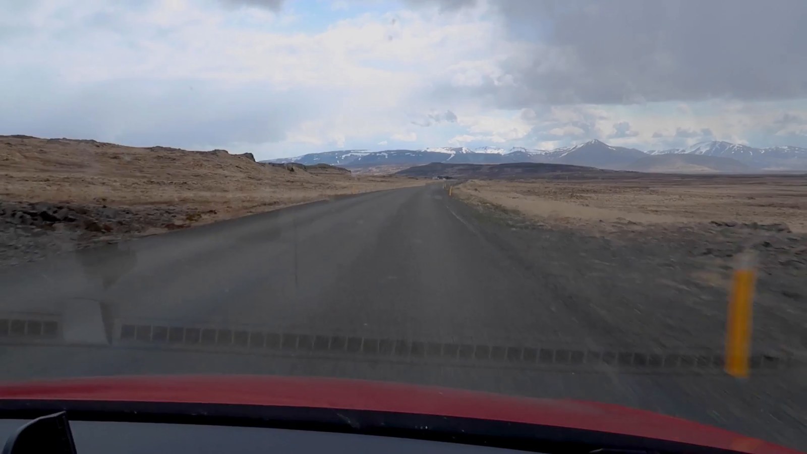 A grey rural road stretching ahead toward distant snow-capped mountains, seen through the windshield of the van.
