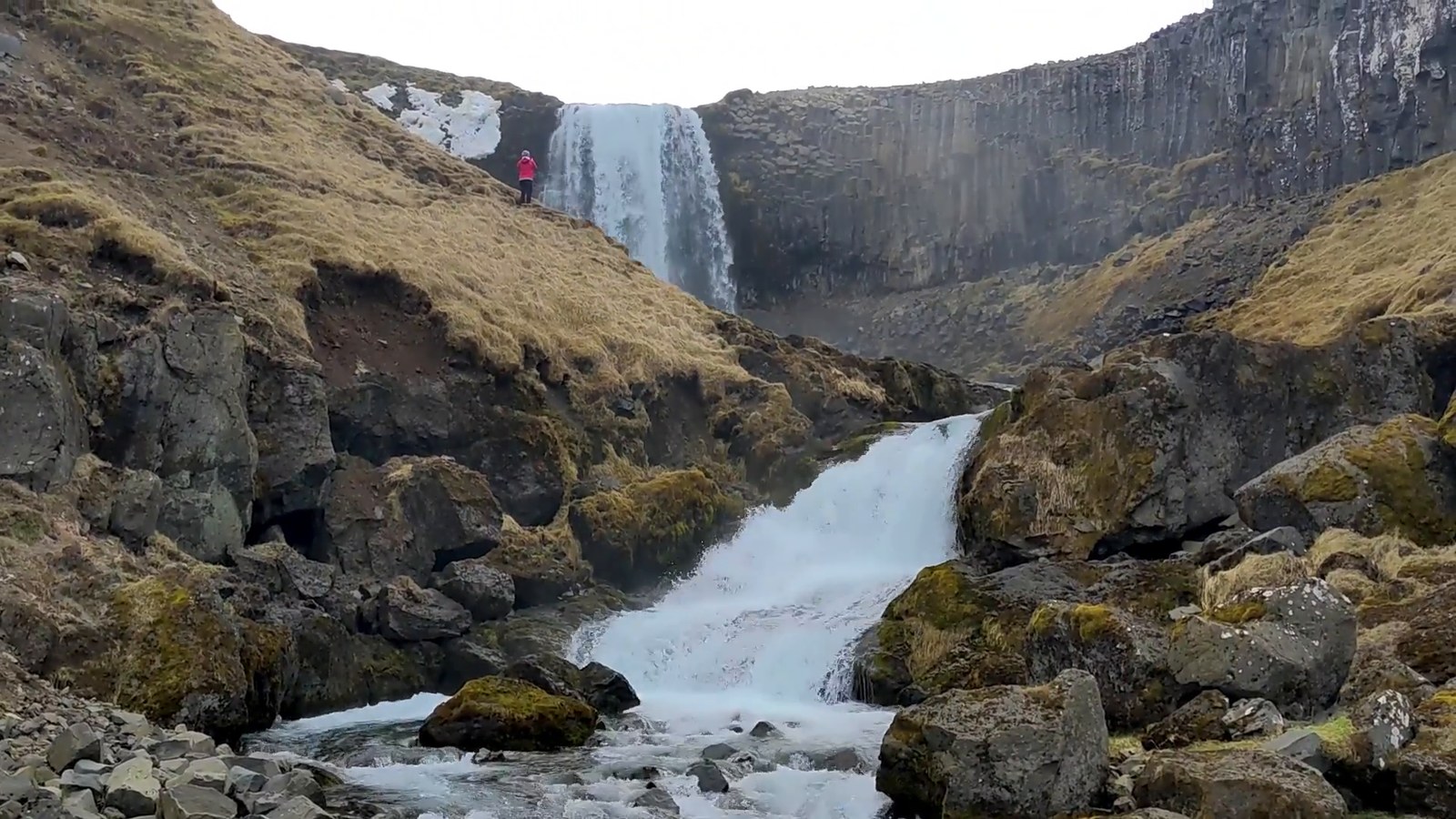 A waterfall dropping over dark basalt cliffs into a rocky stream, with a tiny hiker in a red jacket on the slope.