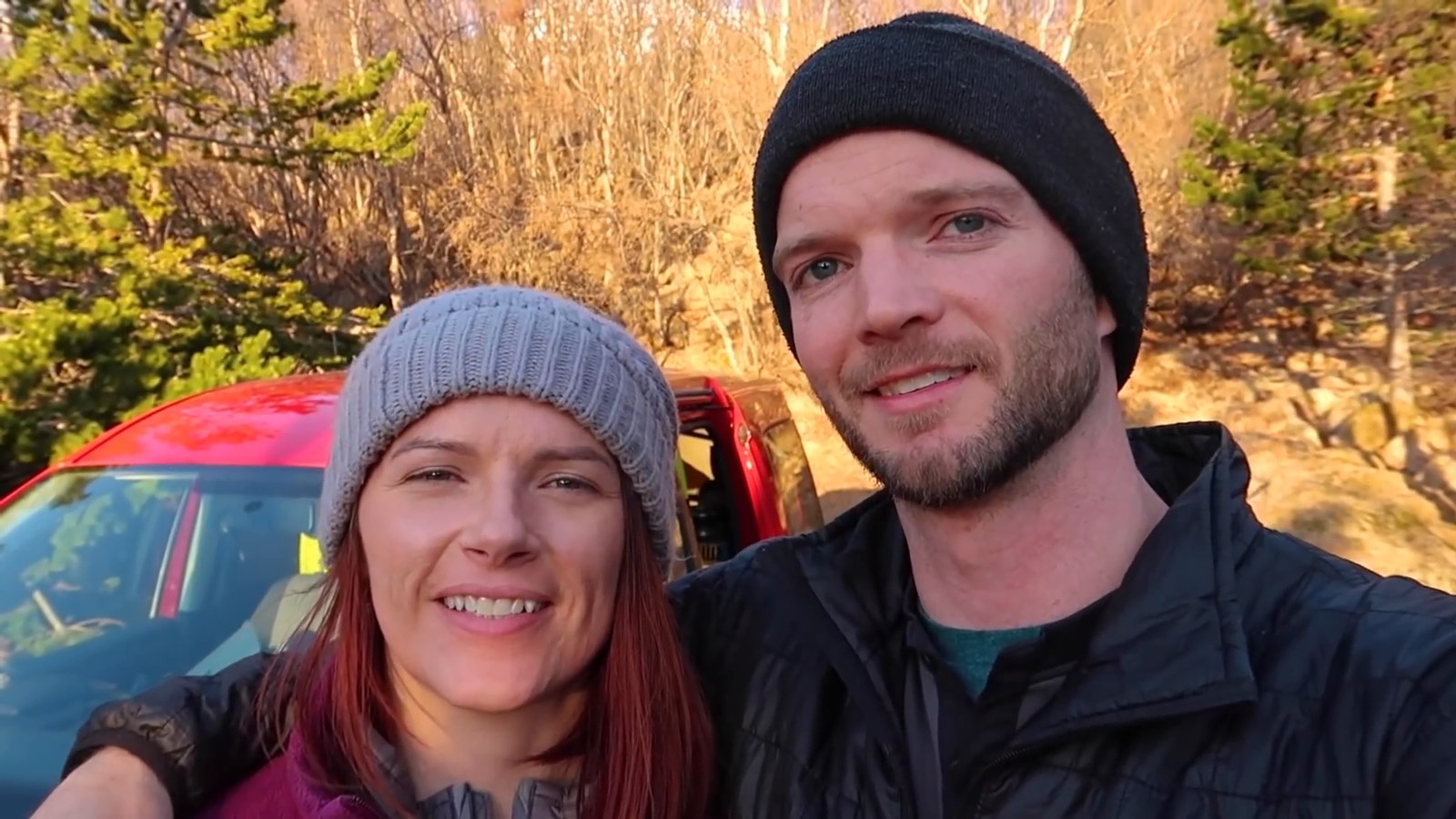 Two people smiling in a selfie in front of a red campervan, with bare golden-brown trees and evergreens behind them in evening light.