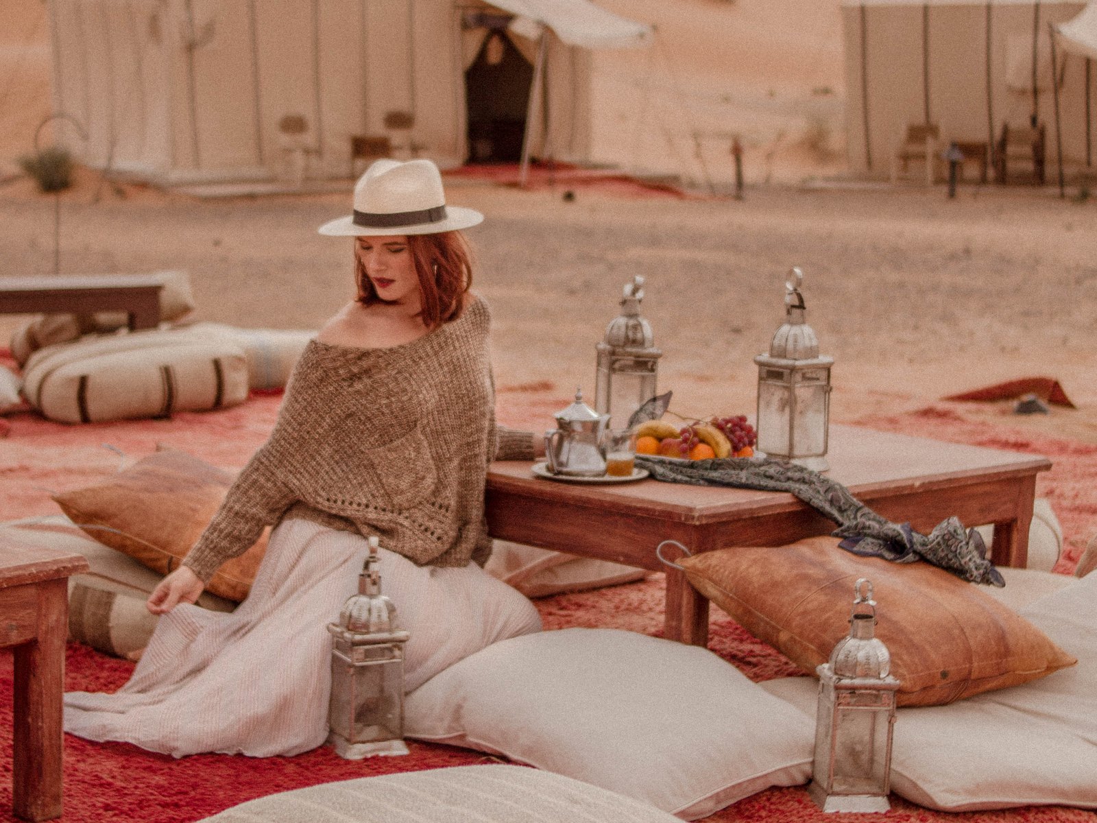 A woman in a straw hat sits on cushions at a low table with tea, lanterns and fruit outside a canvas tent at a desert camp.