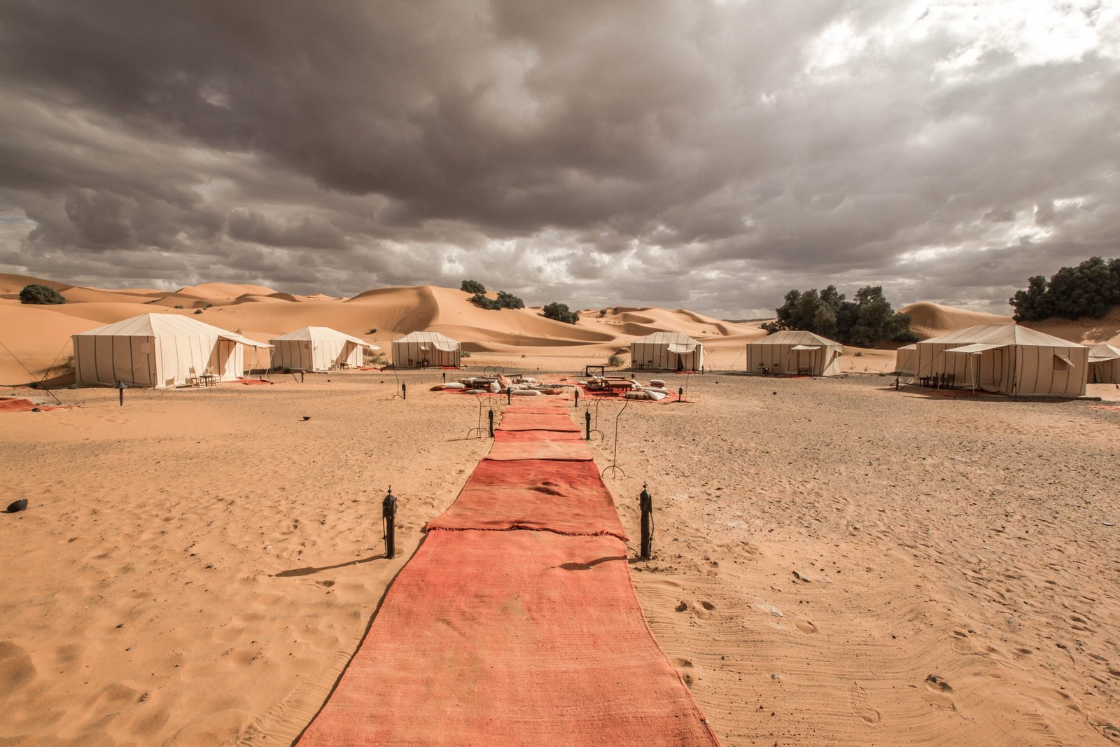 White canvas tents in the dunes at a Merzouga desert camp, with a red carpet leading to a fire-pit area under a stormy sky.