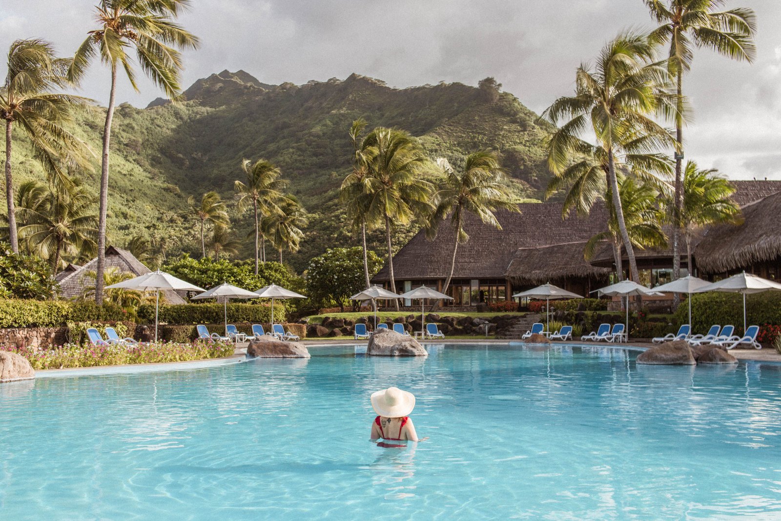 A swimmer in a sun hat stands in a blue resort pool with green mountain peaks and thatched buildings behind it at the Hilton Moorea.