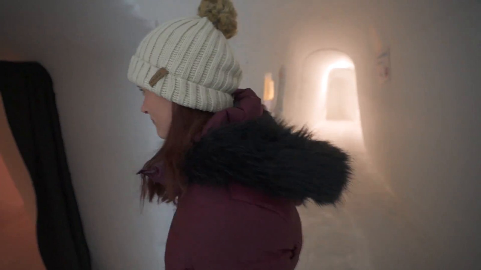 A woman in a white beanie and maroon coat with a fur-trimmed hood walking through a glowing ice-carved corridor.