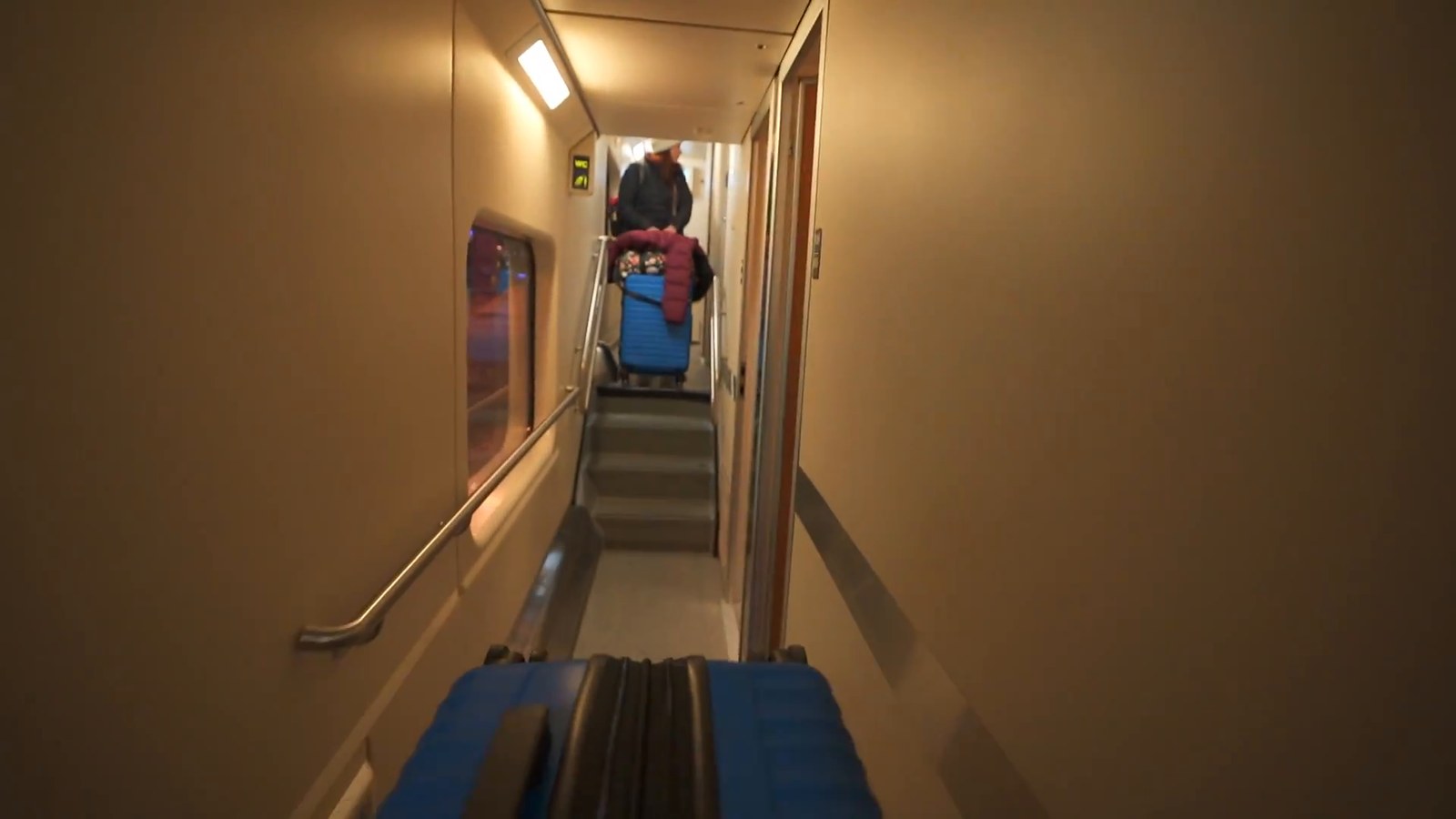 A narrow train corridor with wood-toned walls, a handrail, and a rolling suitcase partway down near a person walking away.