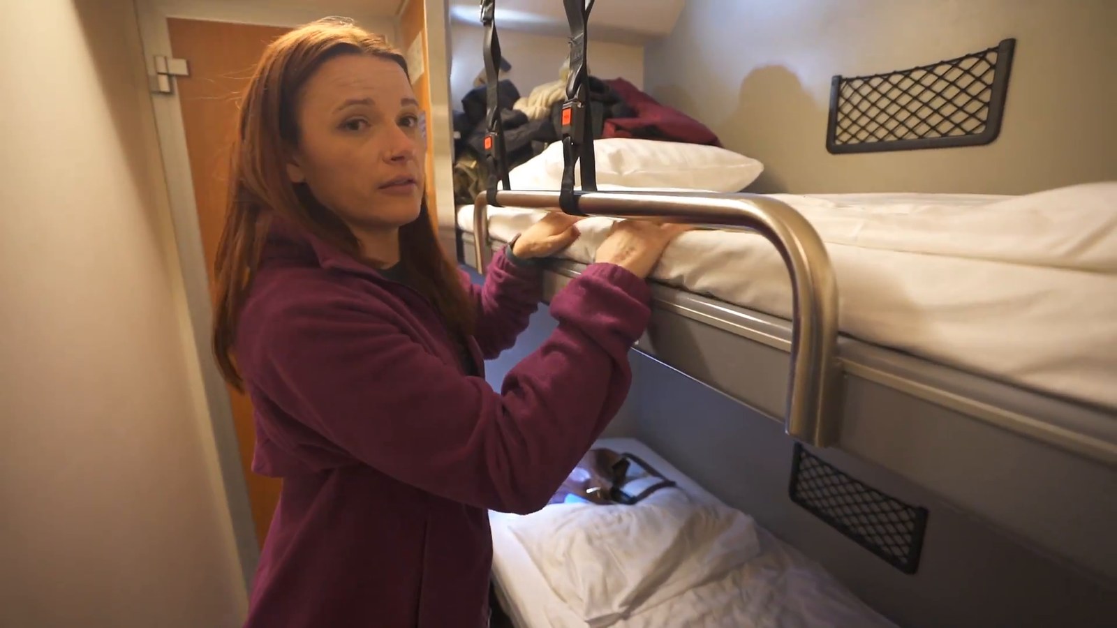 A woman in a maroon fleece adjusting a gear net above a made-up bunk bed with a white duvet and pillow in a train sleeper cabin.