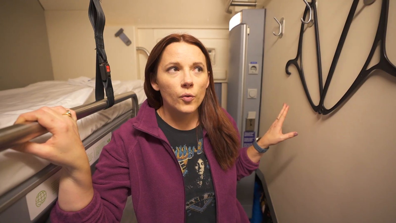 A woman standing in a train sleeper cabin next to a small closet area with hangers and a wall panel with plug sockets.