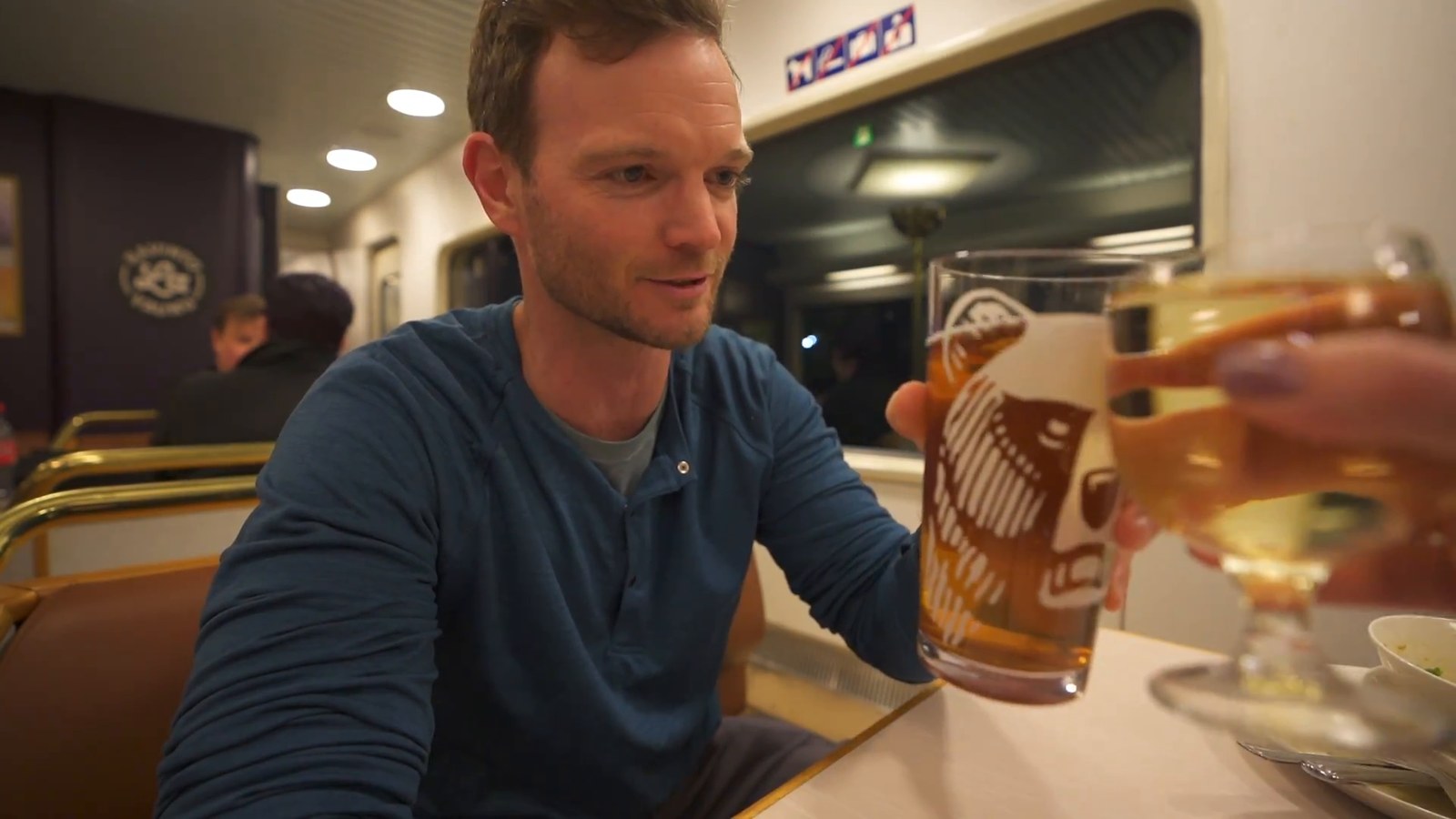 A man in a blue shirt raising a glass of beer in a toast inside a train restaurant car, with dim warm lighting and other seated passengers blurred in the background.