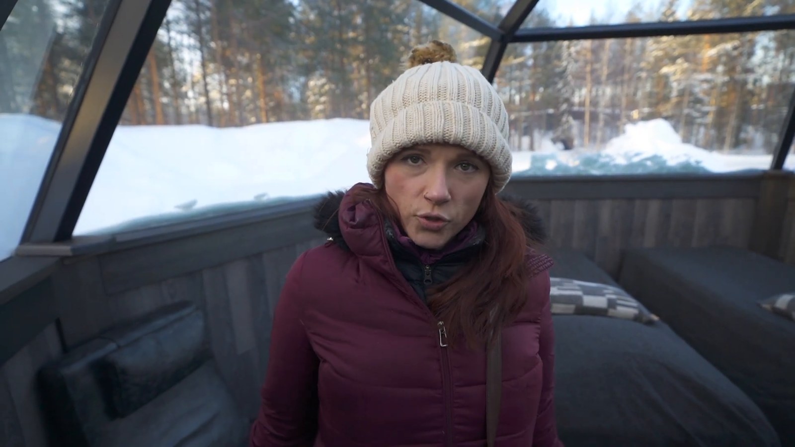 A woman in a white beanie sitting inside a glass-roofed igloo, with snow-covered trees visible through the curved glass structure.