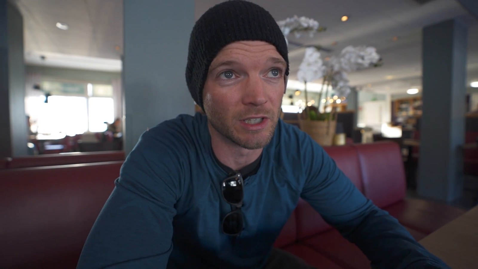 A man in a black beanie sitting at a table in a small station restaurant, with red upholstered booths and a bright window behind him.