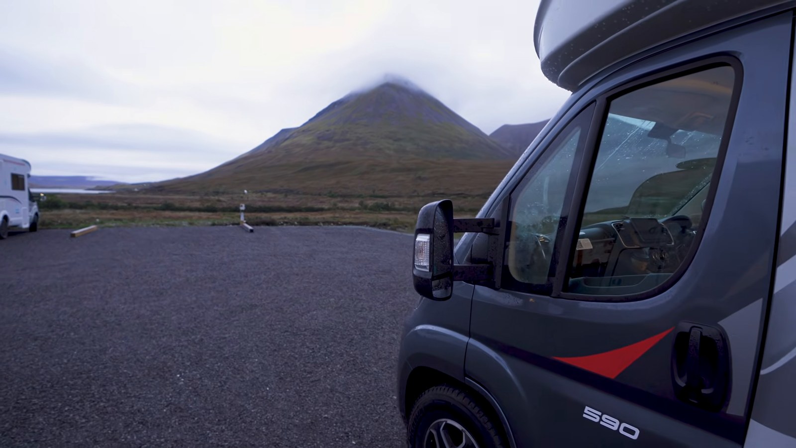 The front of a grey motorhome parked on gravel with a cloud-topped mountain behind, on the Isle of Skye.