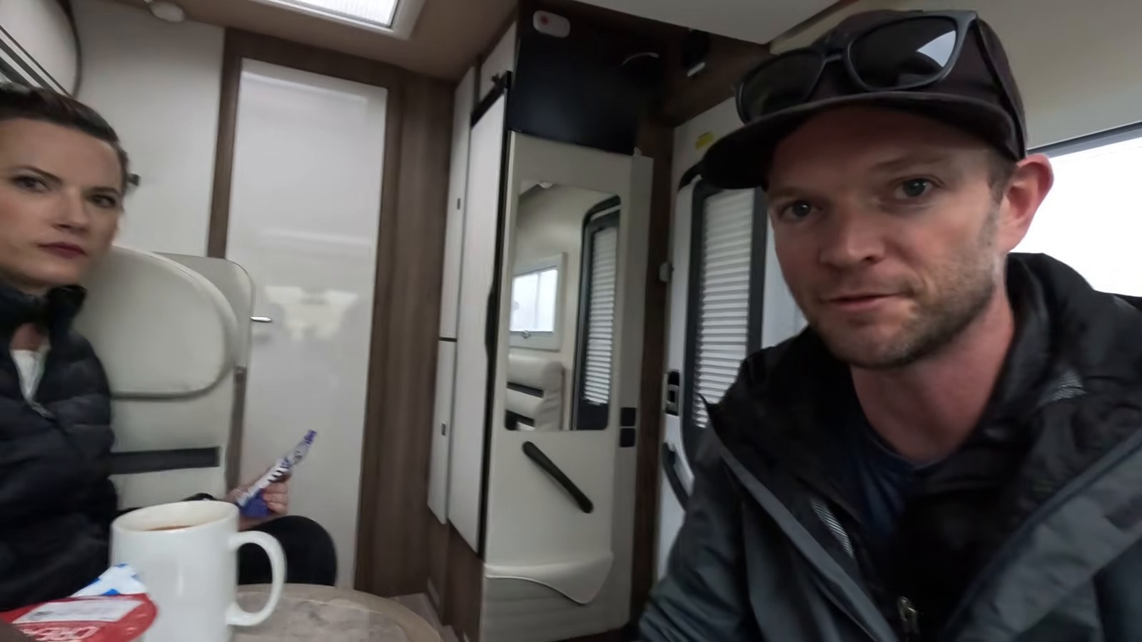 A man and a woman in jackets sitting inside a motorhome with mugs of coffee on the table.