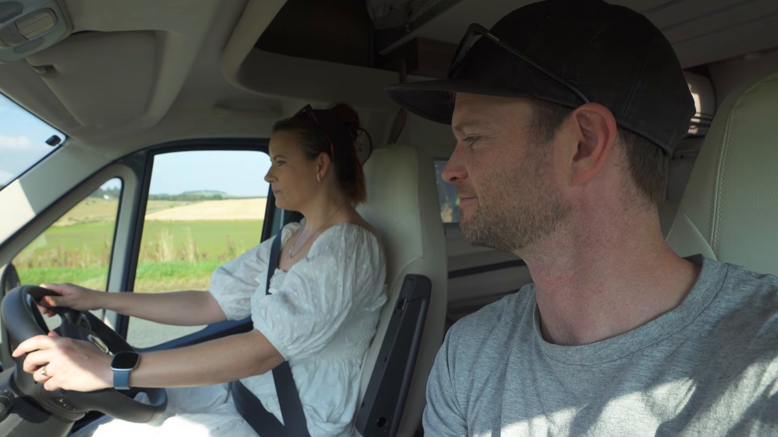 A woman driving a right-hand-drive motorhome along a country road past fields, with a man in a cap in the passenger seat.