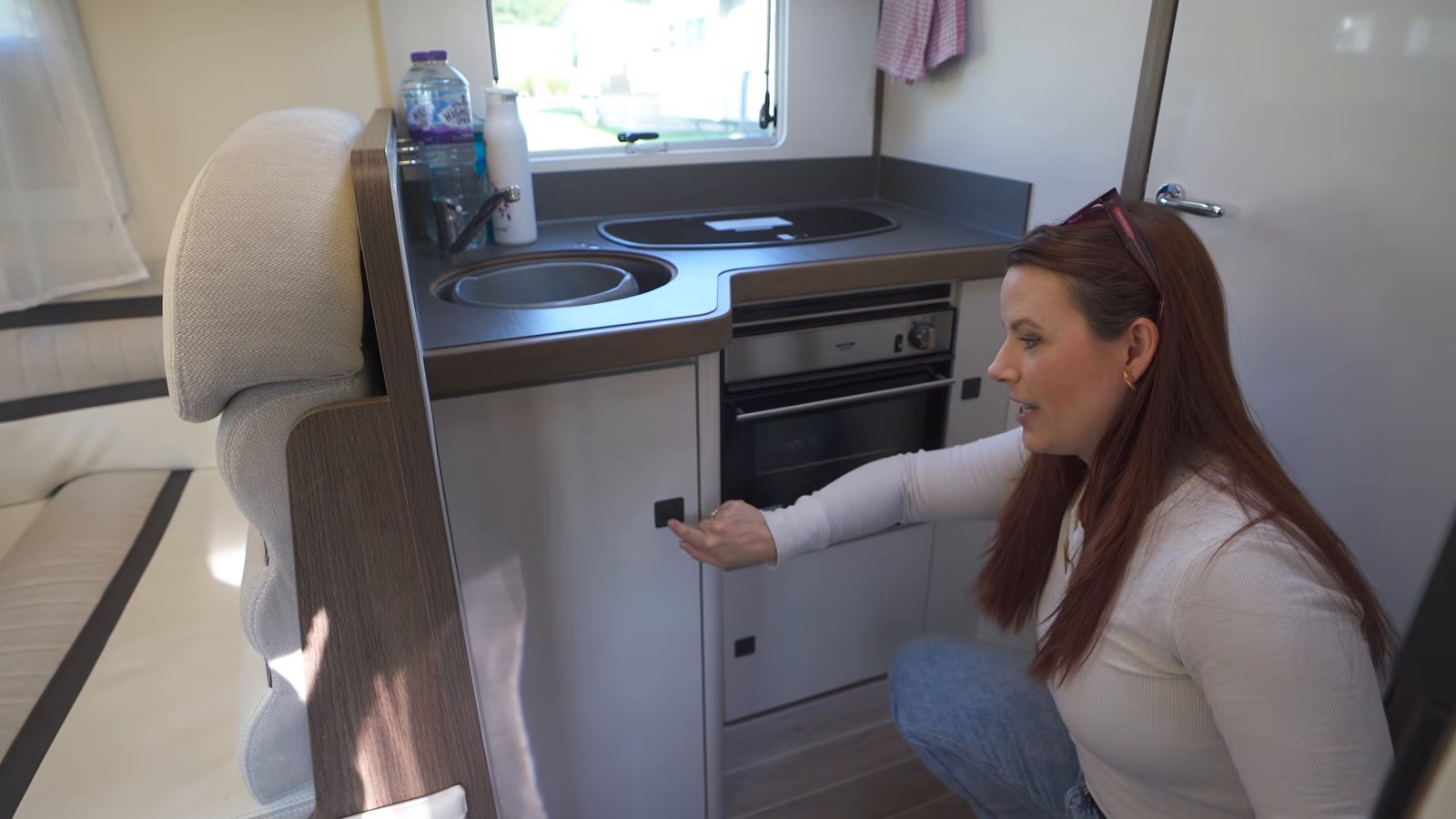 A woman crouching to open the small oven beneath a sink and gas hob in a motorhome kitchen.