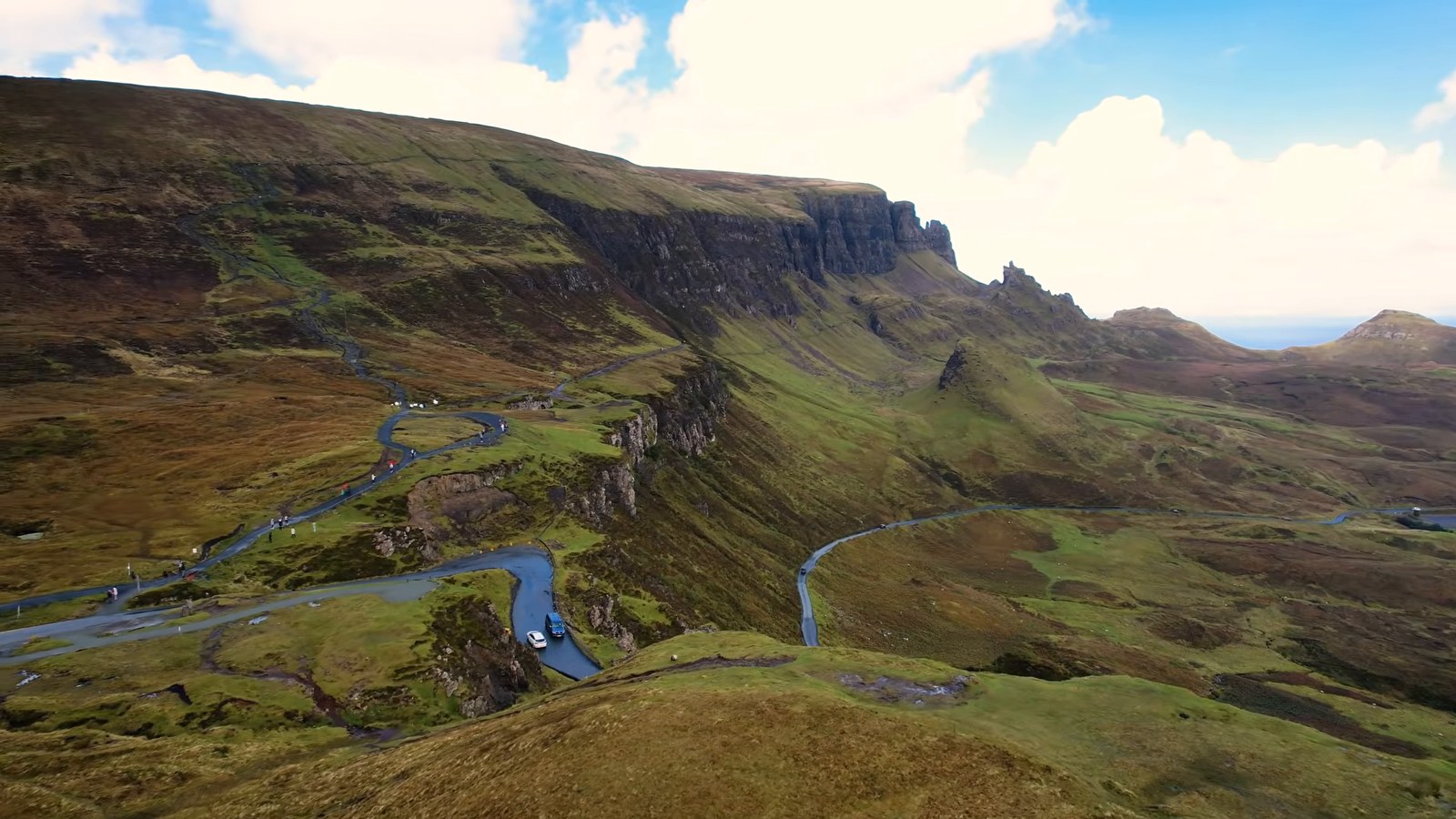 An aerial view of the Quiraing on the Isle of Skye: green slopes and cliffs with a narrow road winding across the valley below.