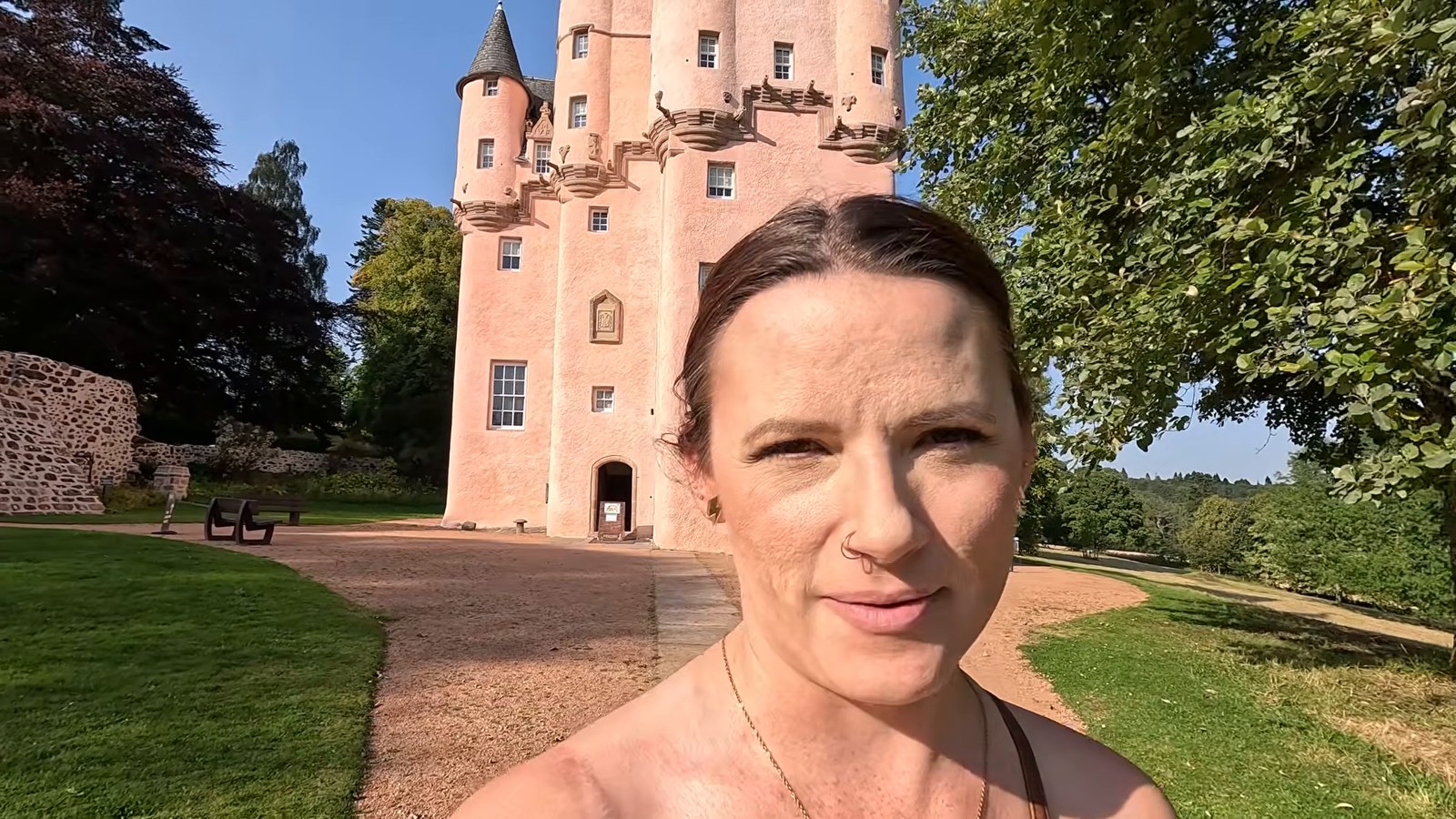 A woman smiling in front of a tall pink tower house with turrets, on a sunny day.