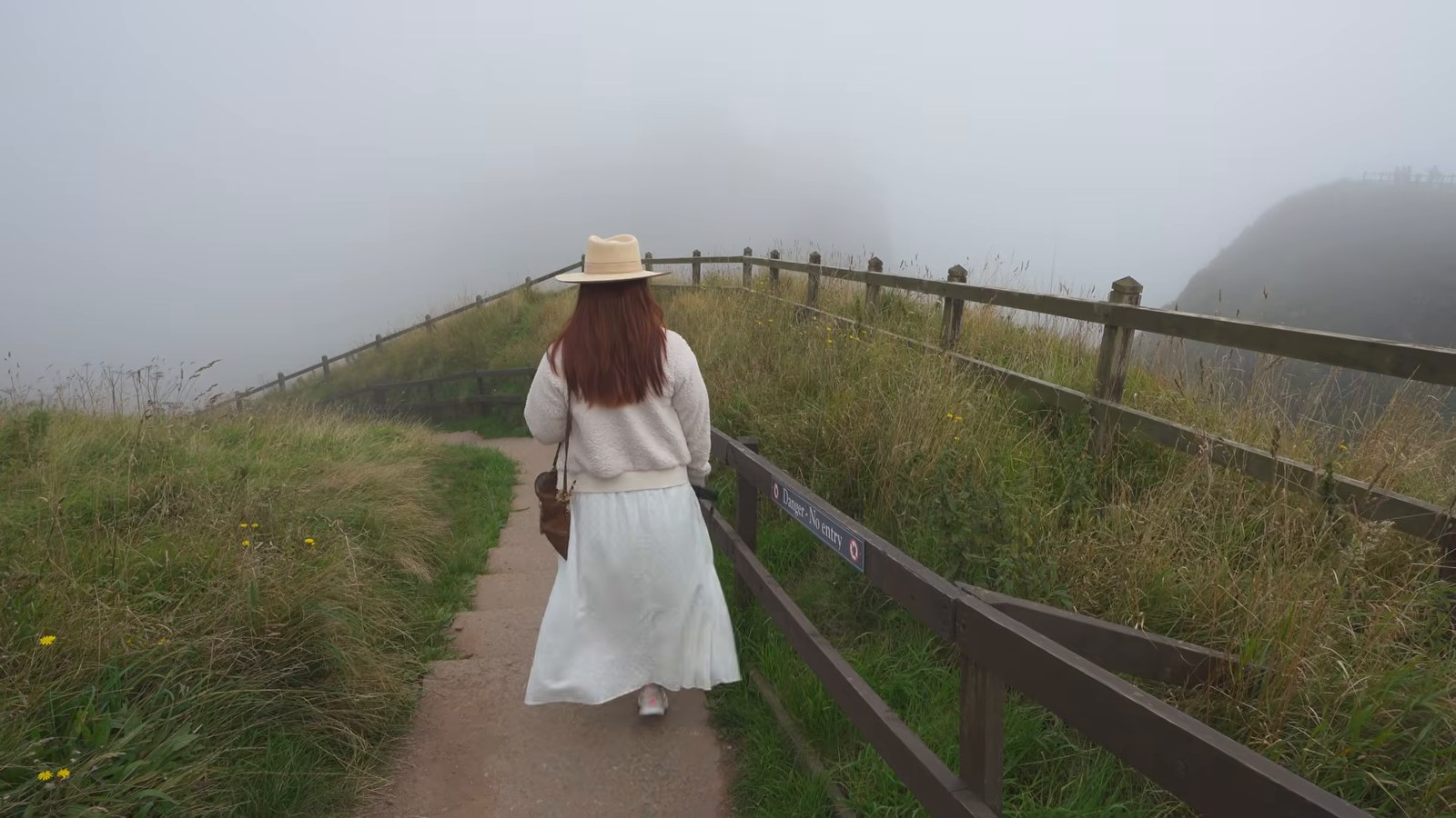 A woman in a white skirt and hat walking along a fenced cliff path into thick fog.