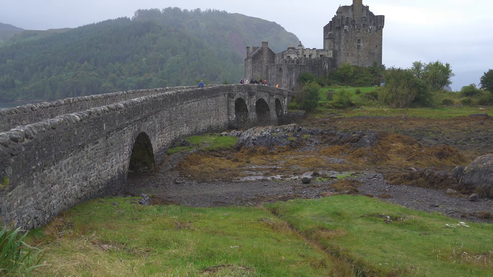 An old stone arched bridge leading across seaweed-covered shore to Eilean Donan Castle under grey skies.