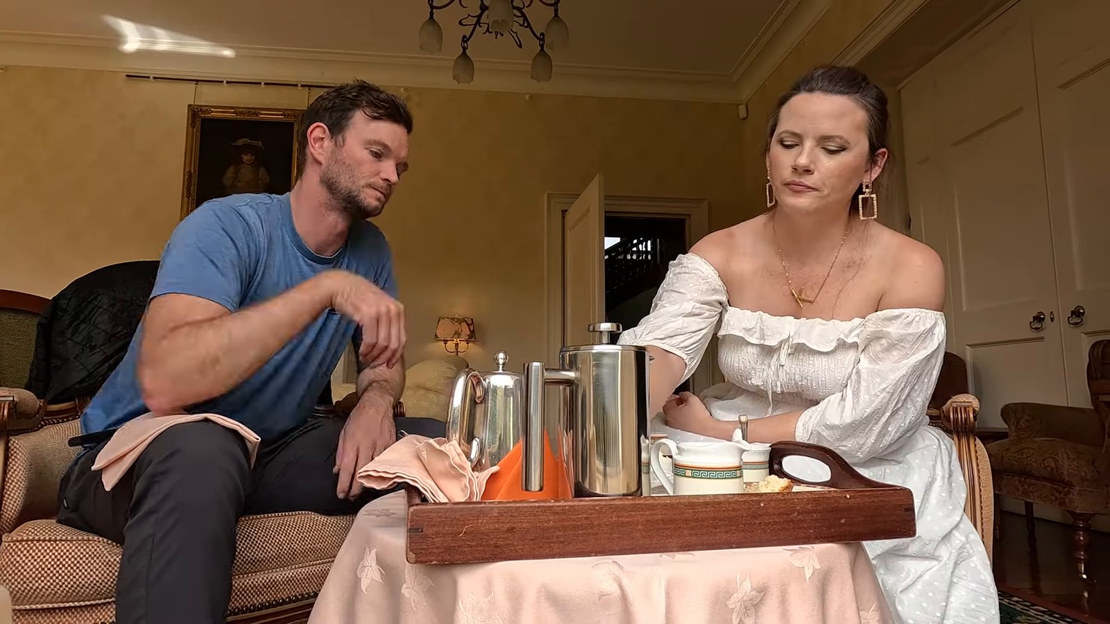 A man and a woman sitting in armchairs in a traditional hotel lounge with a tray of tea things and silver pots.