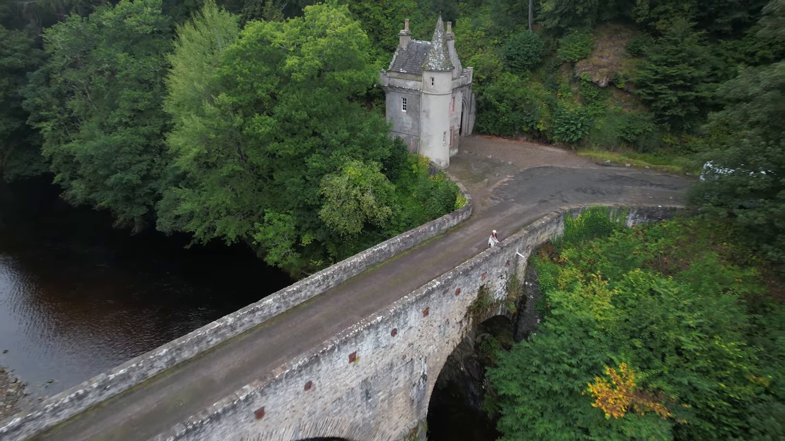 An aerial view of a small turreted gatehouse beside an old stone bridge over a river, surrounded by trees.