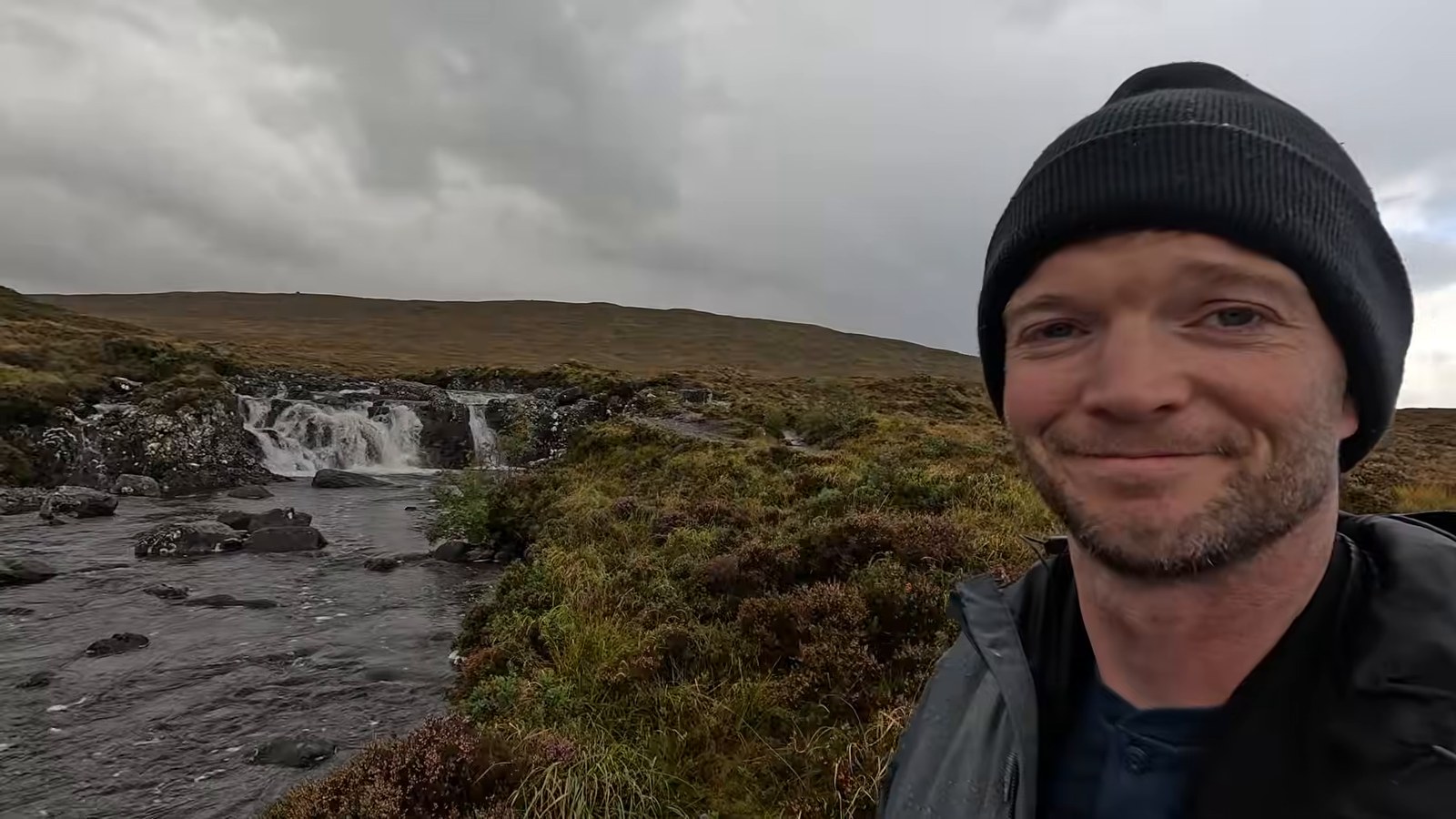 A man in a black beanie smiling beside a small waterfall on a moorland river under a grey sky.