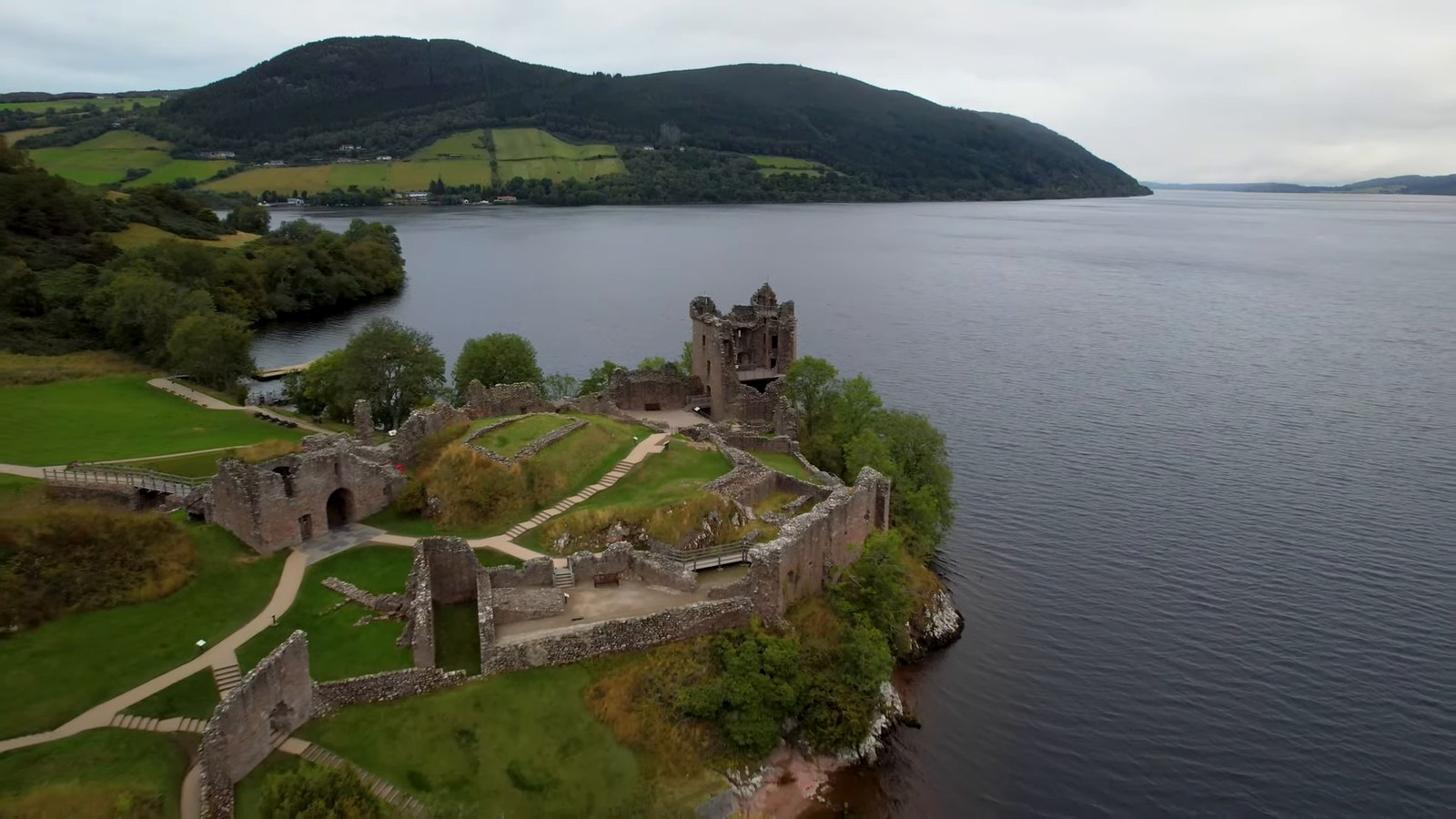 An aerial view of Urquhart Castle's ruins on a green headland jutting into Loch Ness, with hills behind.