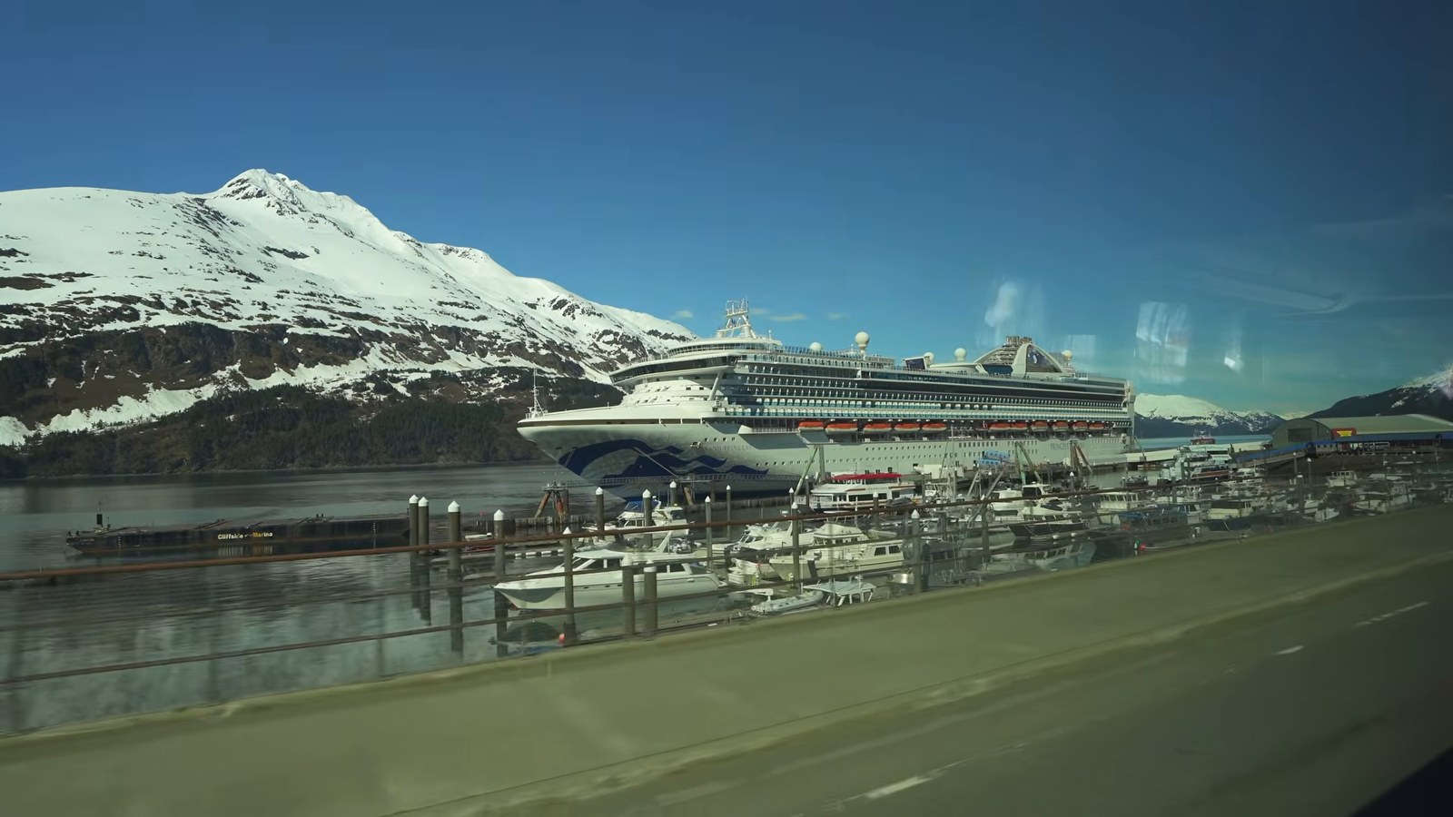 A large white cruise ship docked at a small port town with snow-capped mountains behind it.