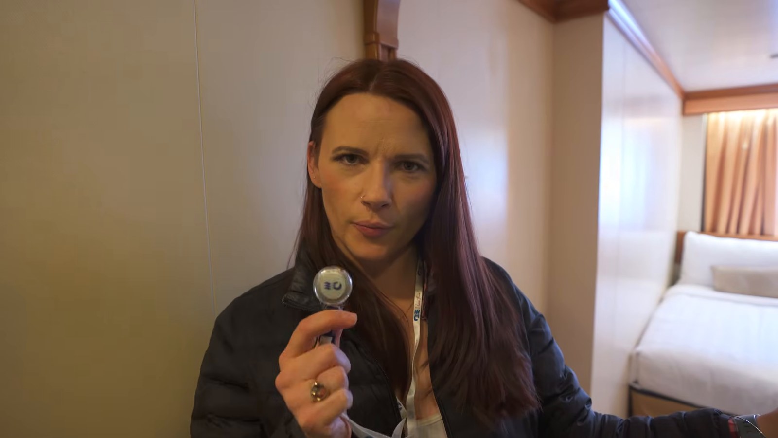 A woman inside a cruise ship cabin holding up a small round medallion toward the camera, with a made-up bed visible behind her.