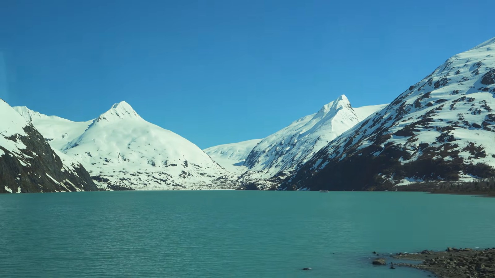 A narrow fjord with calm turquoise water between steep, snow-dusted mountains, seen from a moving vehicle.