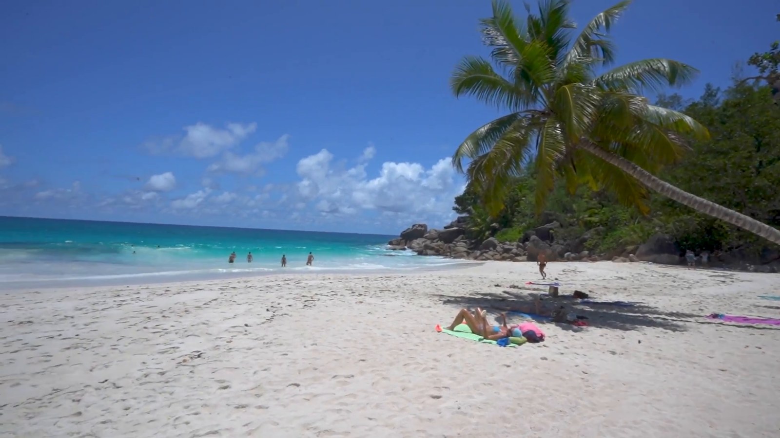 A wide white-sand beach with turquoise water, a leaning palm and granite boulders under a blue sky, with a few people far in the distance.