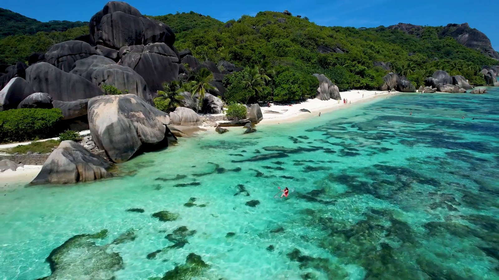 Aerial view of turquoise shallow water and huge granite boulders along a white-sand beach backed by green hills on La Digue.