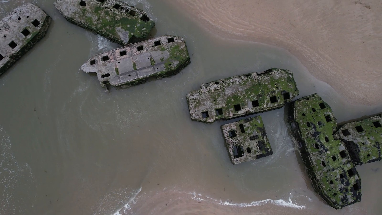 An aerial view of weathered concrete harbor sections scattered in the shallows off the beach at Arromanches.