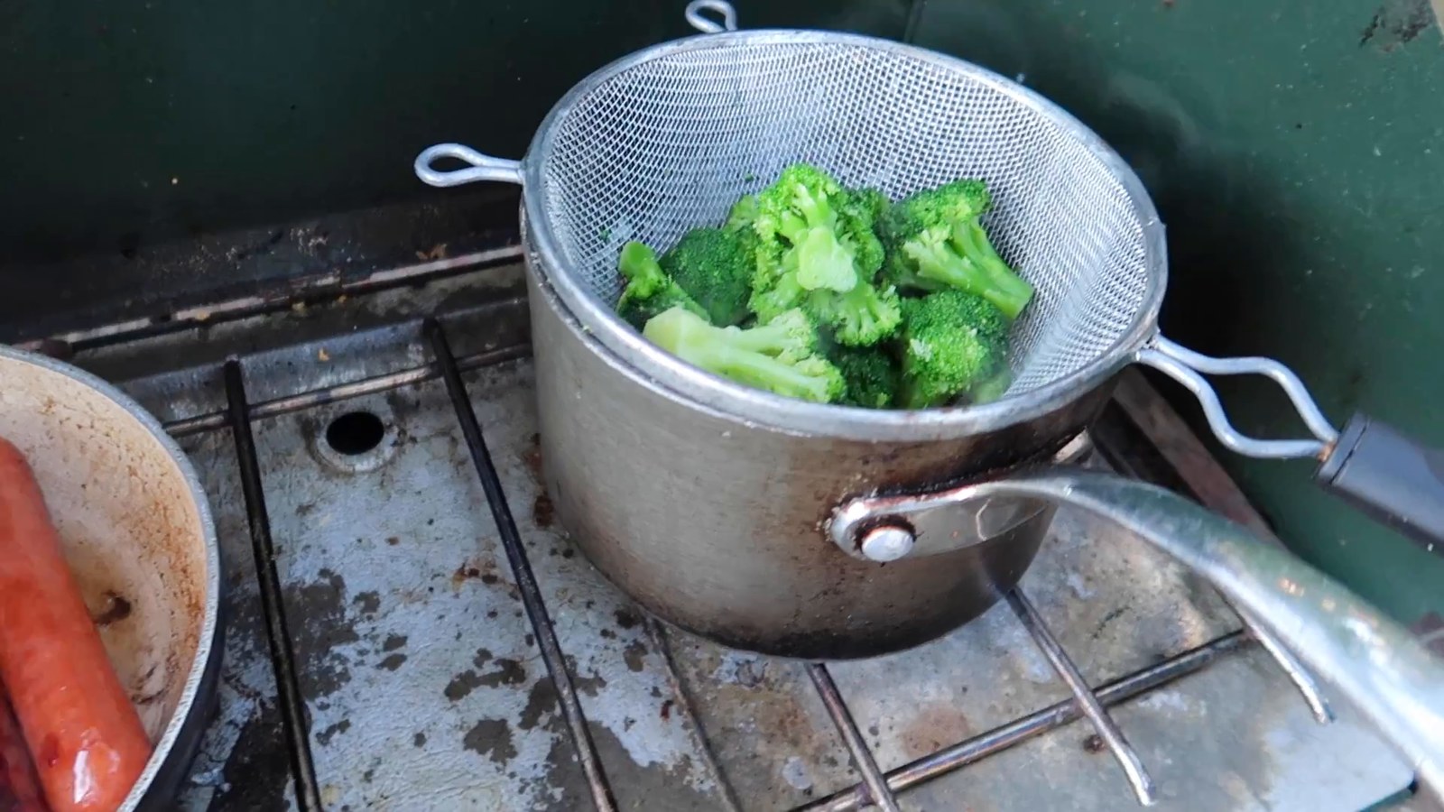 A strainer of broccoli and a pot with a sausage sitting on a camp stove.