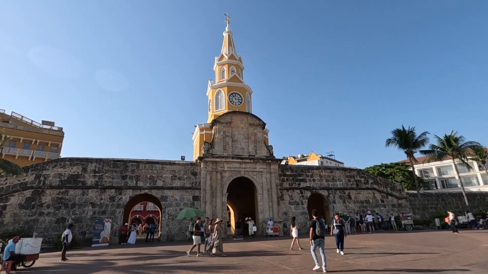 The yellow Clock Tower above the arched gate in Cartagena's old city wall, with a few visitors crossing the plaza under a blue sky.