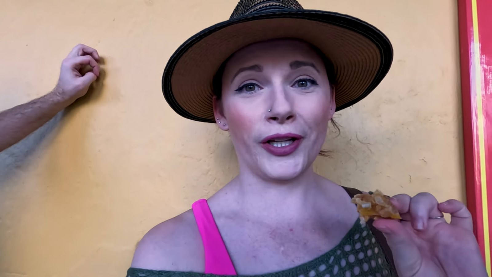 A woman in a straw hat holds up a golden, crisp Colombian snack in front of a mustard-yellow wall.
