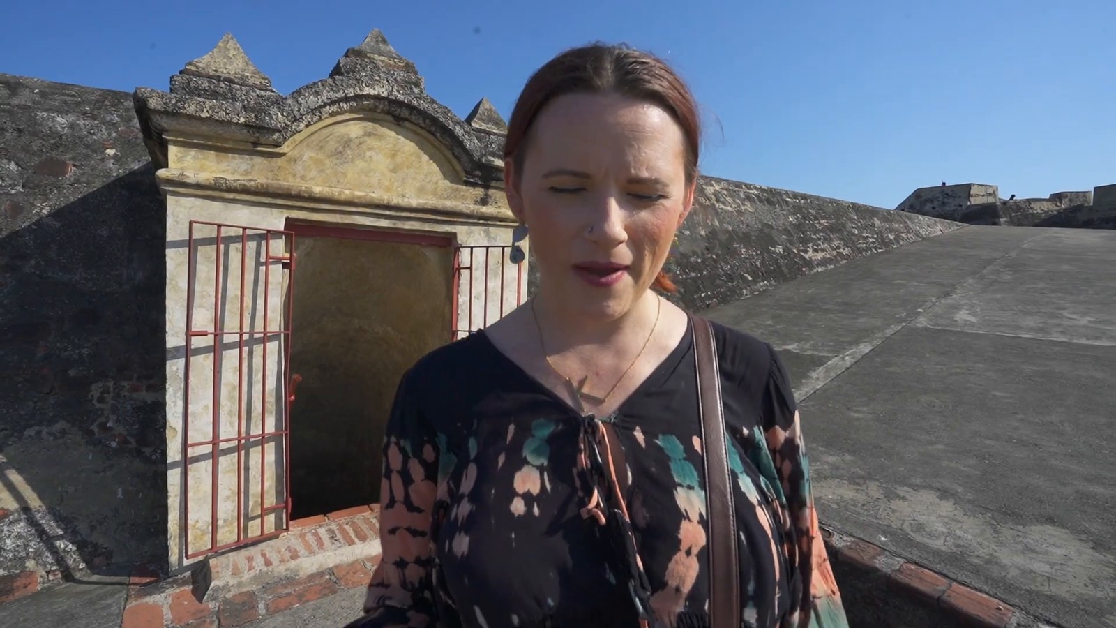 A woman stands beside a small stone alcove with a red iron gate set into the wall at Castillo San Felipe.