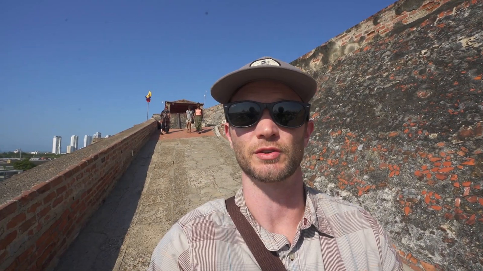 A man in sunglasses and a cap stands on a sunny stone ramp at Castillo San Felipe, with brick and weathered fortress walls beside him and the city skyline in the distance.
