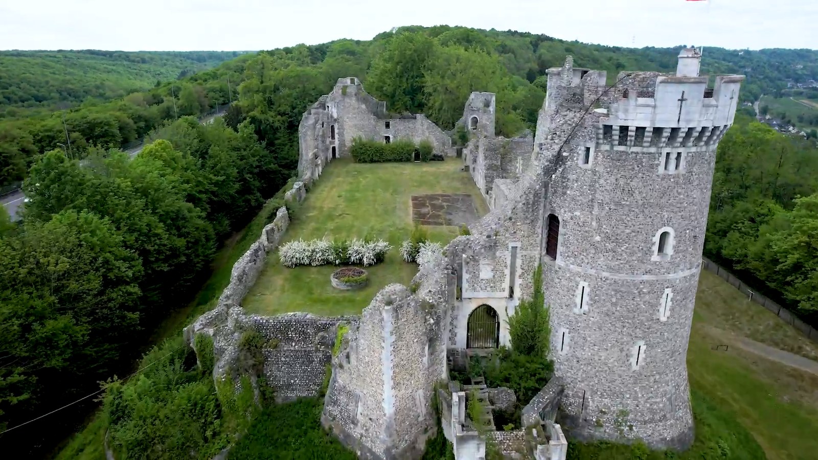 An aerial view of a ruined stone castle with a tall round tower and a grassy courtyard beside a wooded road.