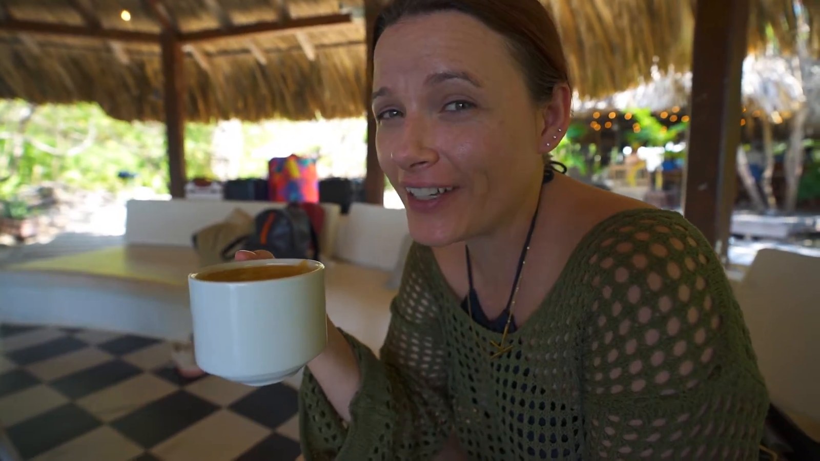 A woman holds up a white cup of coffee under a thatched roof, with a checkered tile floor and a white lounge area behind her.