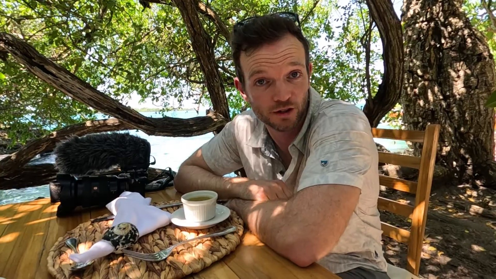 A man rests his arm on a wooden table under tree branches by the water, with a cup of soup, cutlery on a woven mat and a camera beside him.