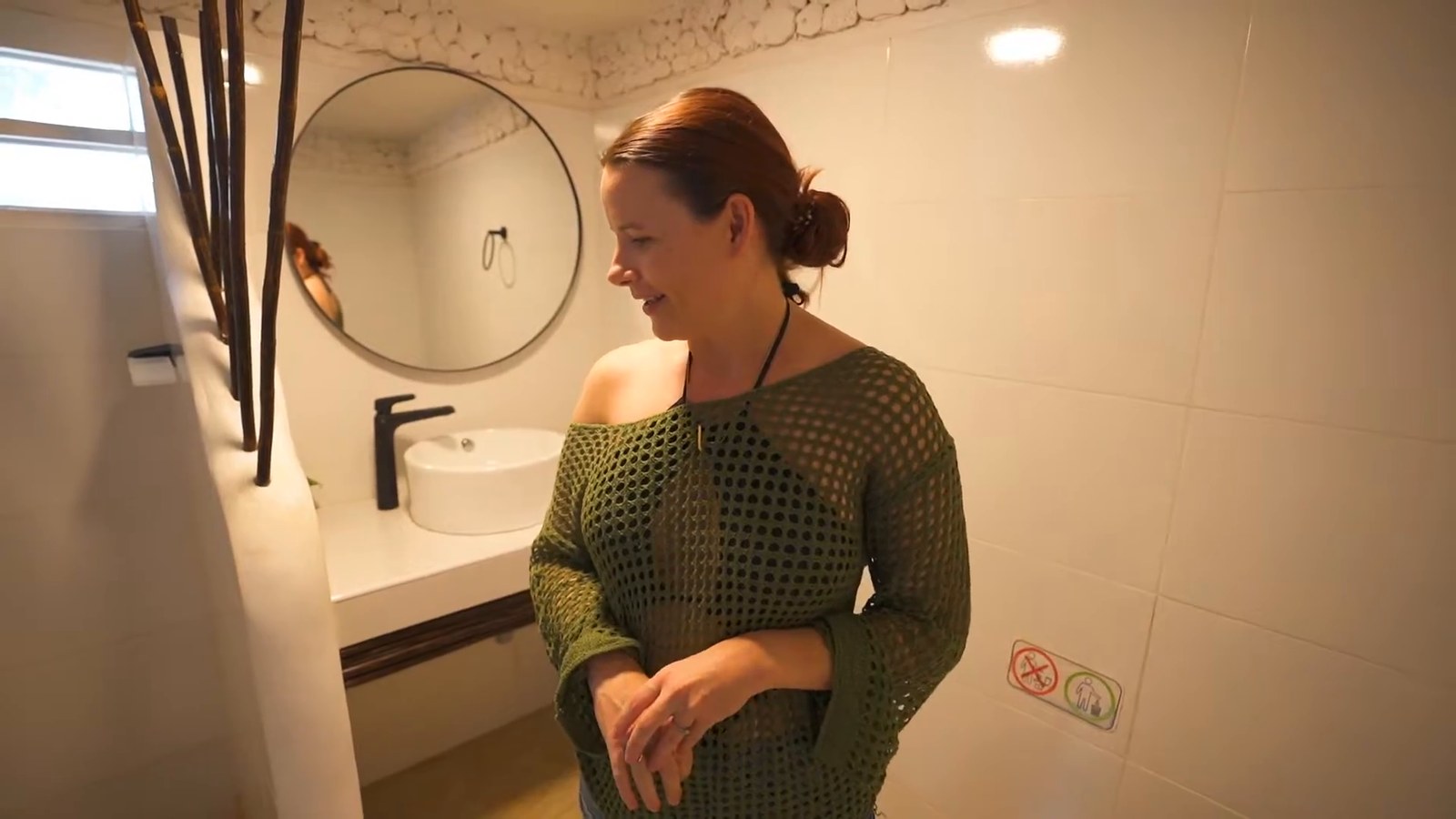 A woman stands in a bright white-tiled bathroom with a round mirror, a black faucet and a white vessel sink.