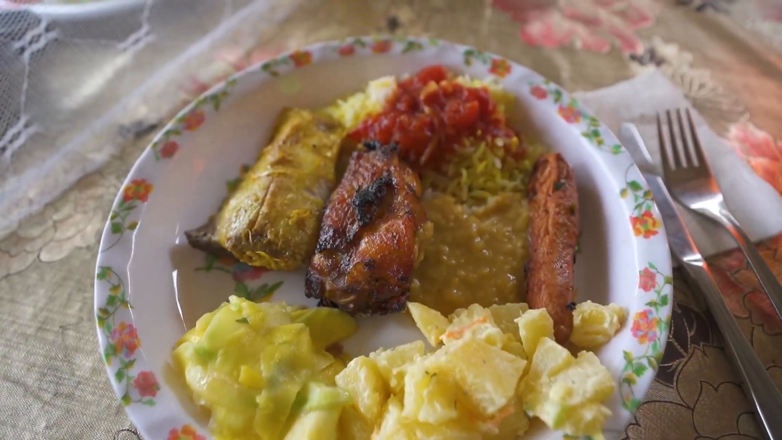 A plate of grilled fish, rice, red creole sauce, lentils and potato salad on a flowered plate with a knife and fork.