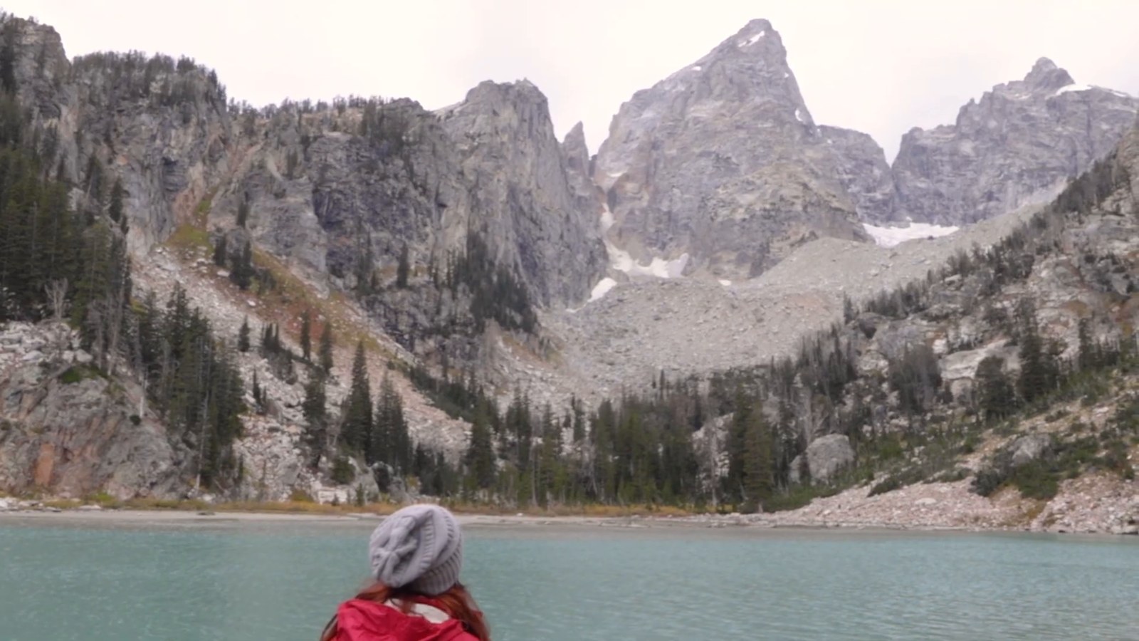 A hiker in a grey beanie and red jacket looks across a turquoise mountain lake toward rugged Teton peaks under a cloudy sky.