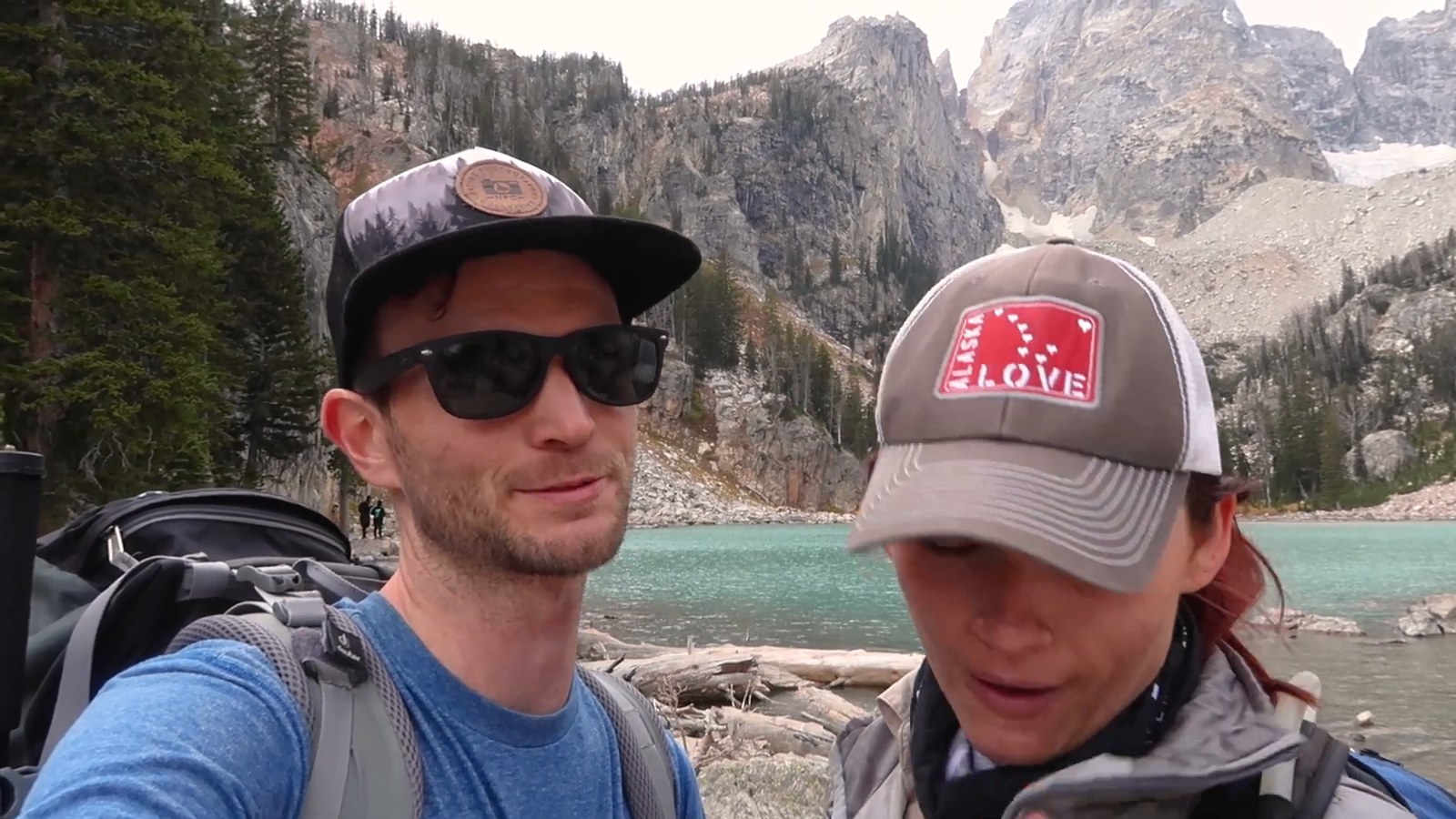 Two hikers in caps and packs pose beside a turquoise lake with Teton peaks rising behind it.