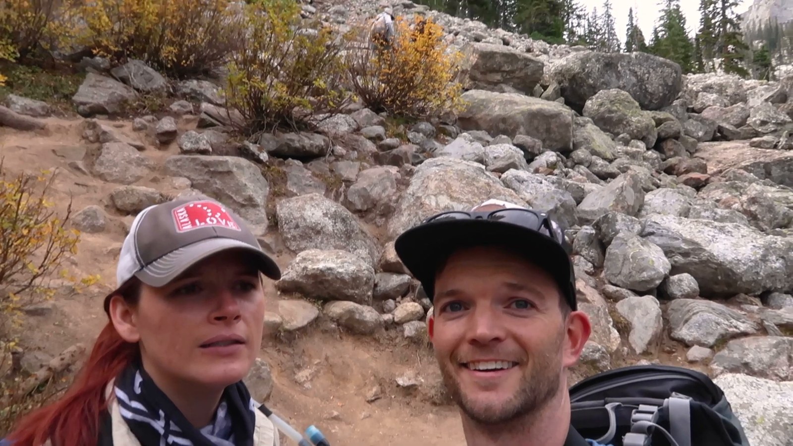 Two hikers in caps smile at the camera in front of a slope of grey boulders on the unmarked Delta Lake route.
