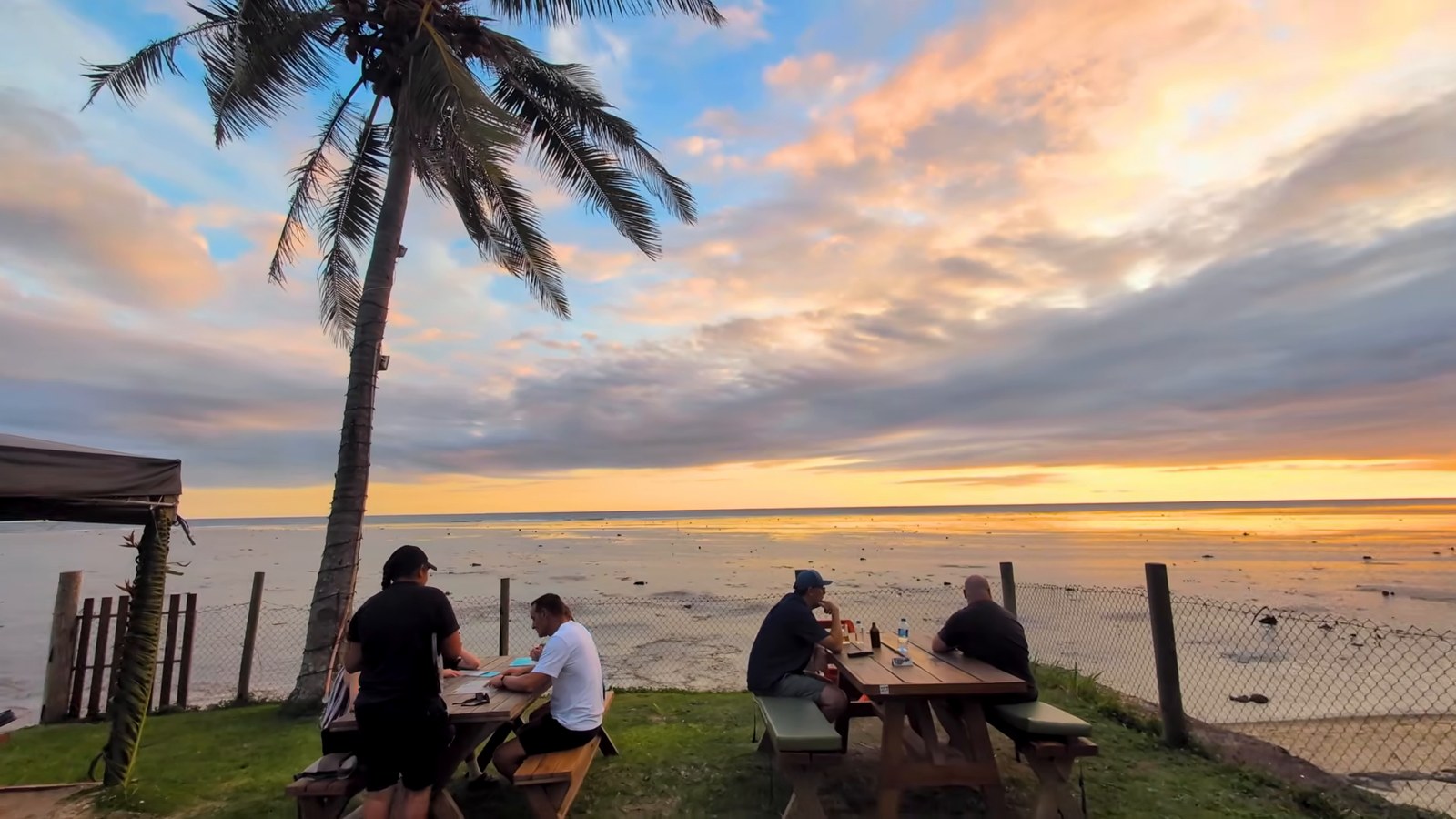 People seated at picnic tables under a palm tree, watching a colorful sunset over a beach on Fiji's Coral Coast.