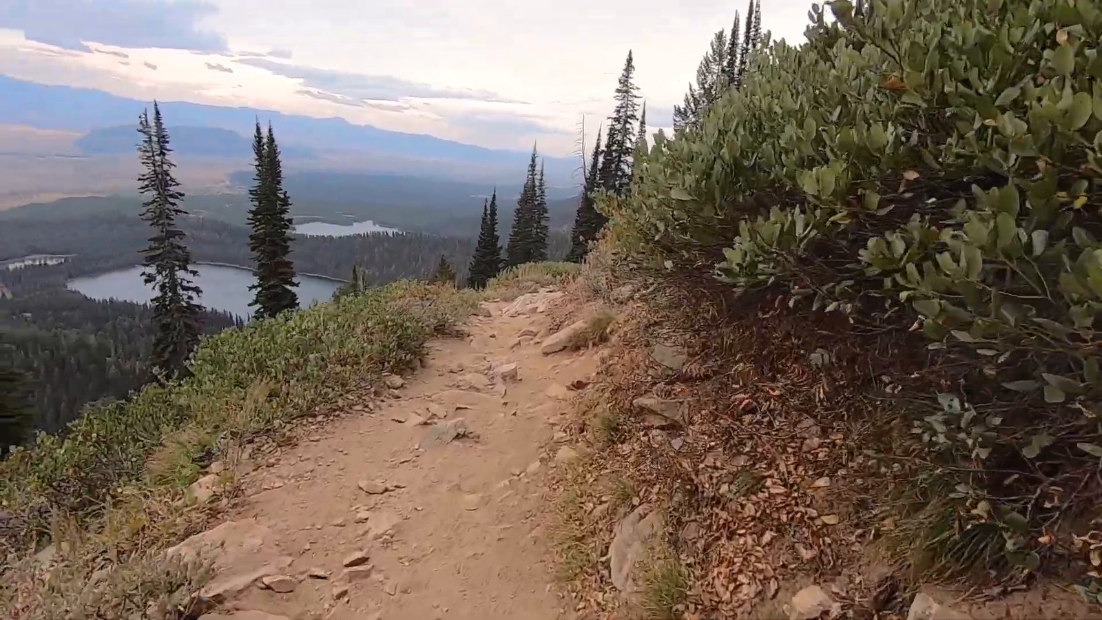 A dirt trail along a hillside shrub with lakes and a forested valley spread out below under a pale sky.