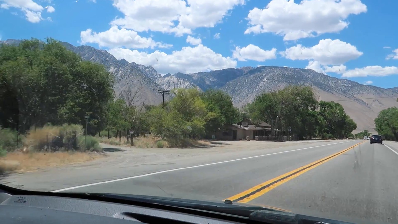 A two-lane highway with a double yellow line running toward mountains under a blue sky with scattered clouds.