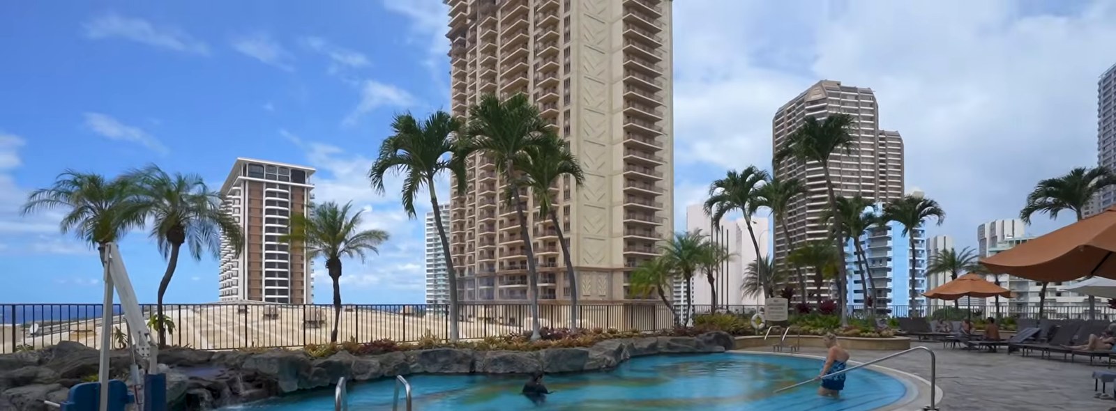 A small rock-lined pool with steel handrails on a stone terrace, with towers and palm trees behind it.