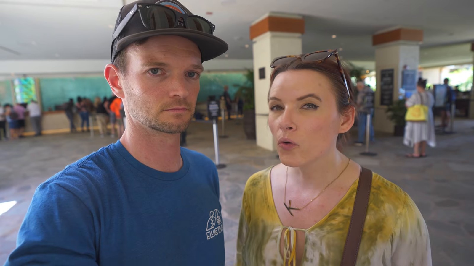 Two travelers take a selfie in the open-air main lobby at Hilton Hawaiian Village, with the long check-in line behind them.