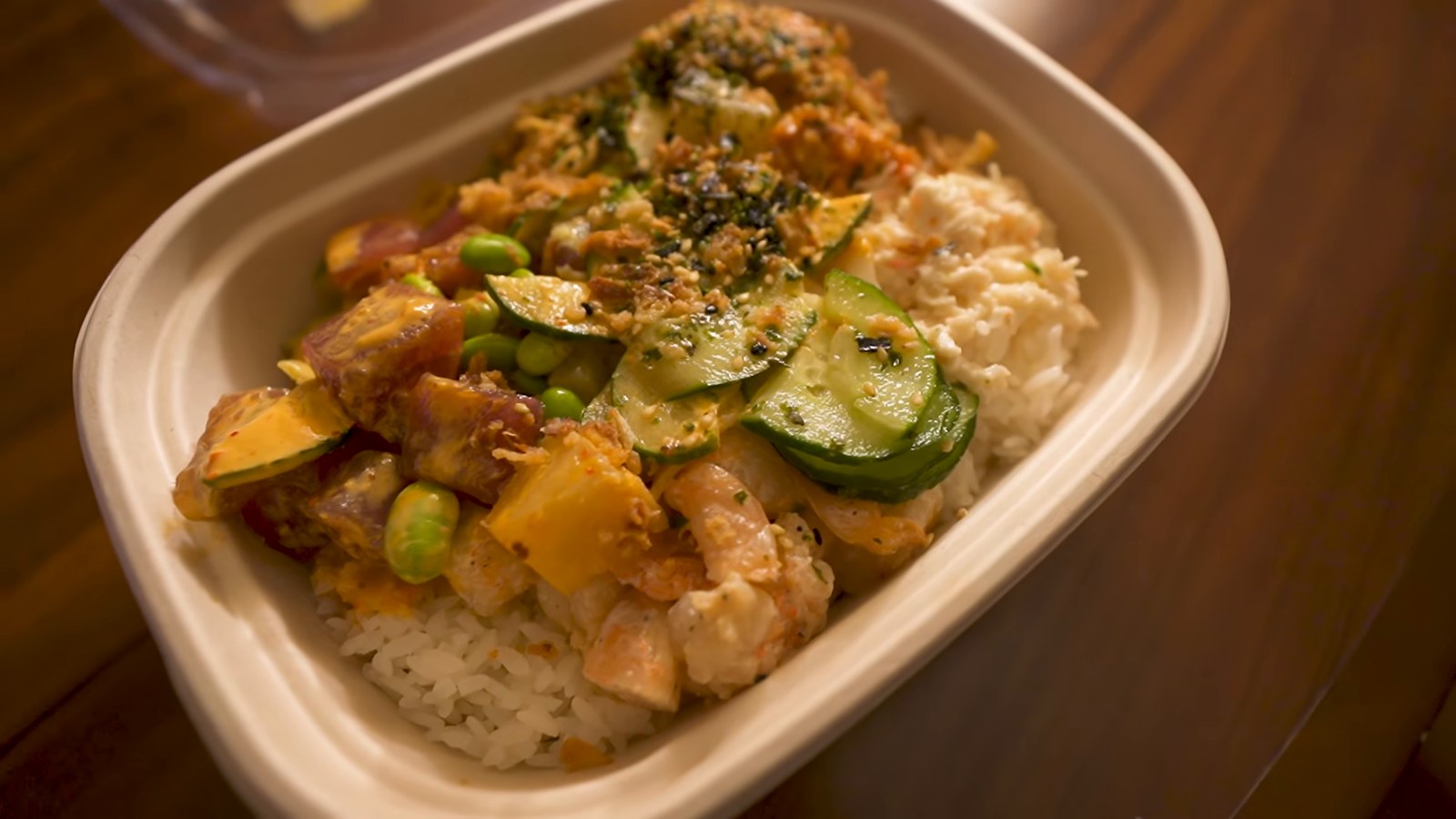 A takeout bowl of poke with rice, cucumber, edamame and diced fish on a wooden table.
